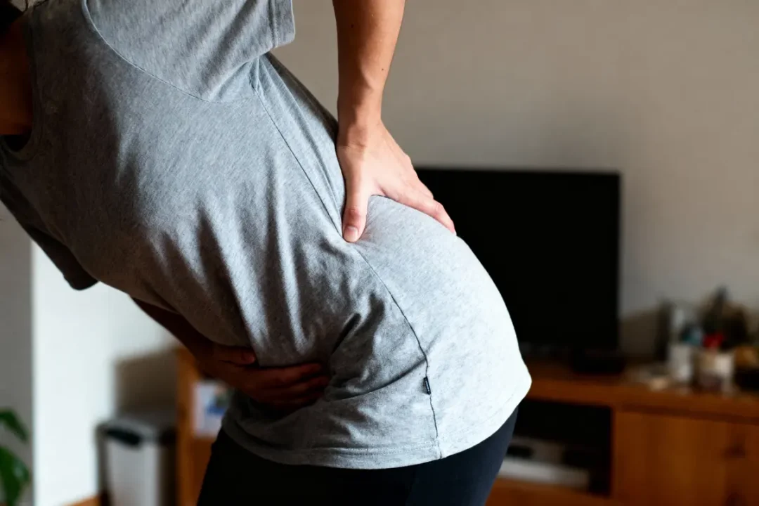 Personne souffrant de douleur au dos, mains posées sur les hanches, portant un t-shirt gris et pantalon noir.