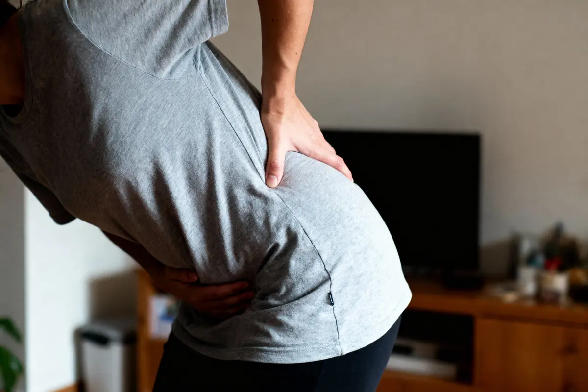 Personne souffrant de douleur au dos, mains posées sur les hanches, portant un t-shirt gris et pantalon noir.