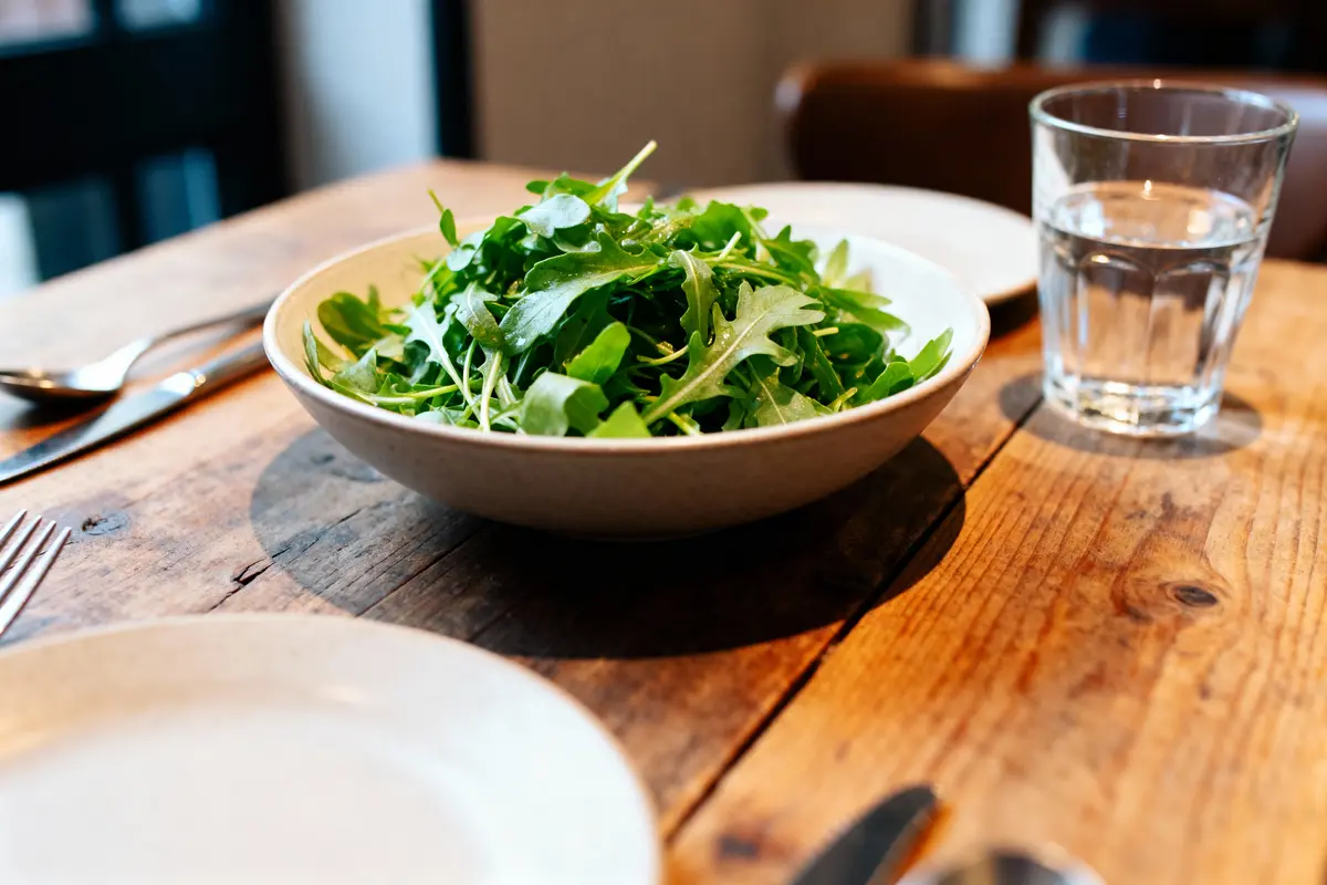 Assiette de roquette fraîche sur table en bois, avec verre d'eau et couverts à côté.