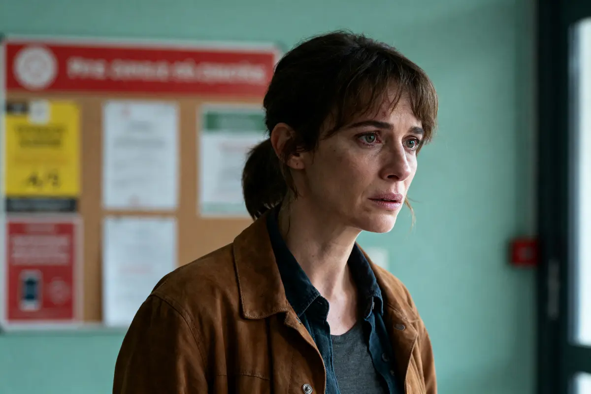 Femme brune au regard sérieux, portant veste marron, fond flou avec panneaux d'affichage multicolores.