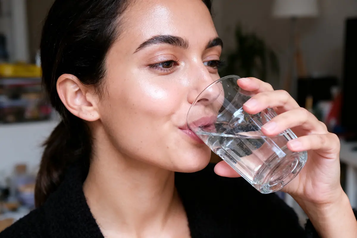 Femme buvant un verre d'eau chez elle, portant un pull noir, ambiance intérieure décontractée.