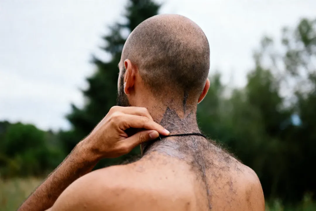 Homme de dos avec cheveux rasés, tenant un collier, tatouage sur la nuque, nature en arrière-plan.