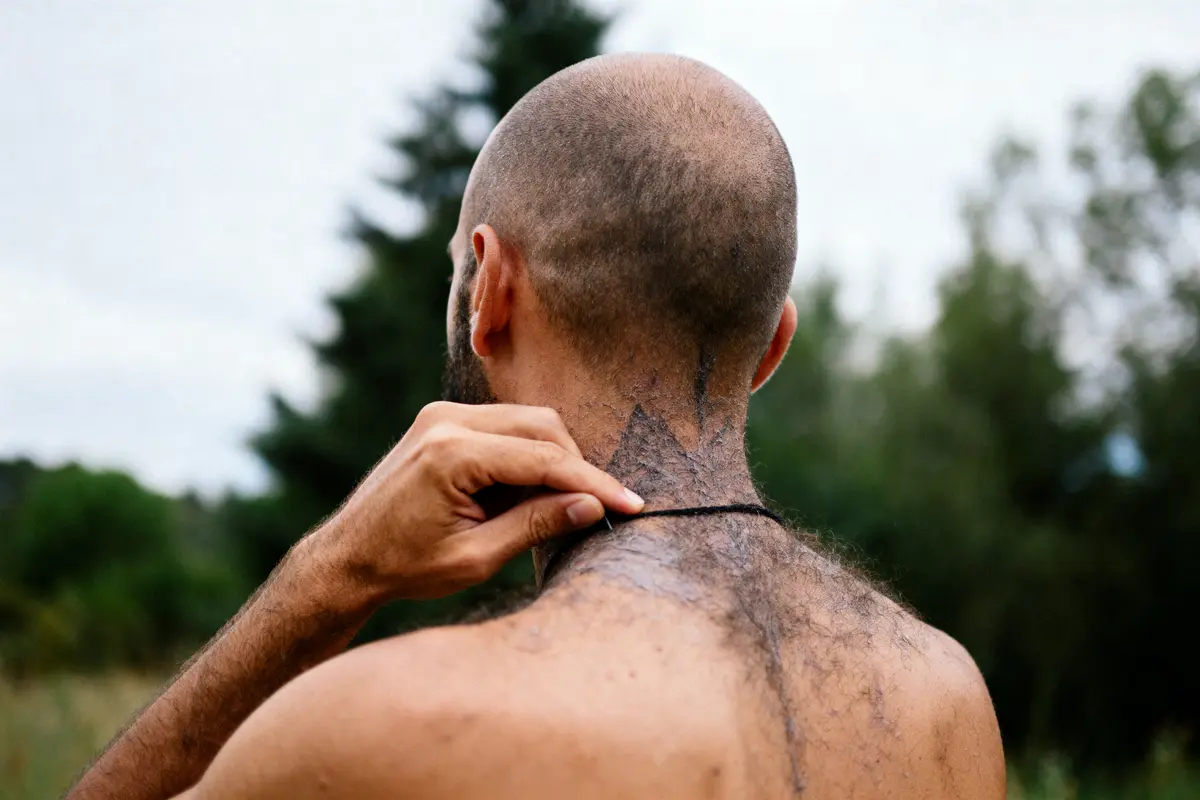 Homme de dos avec cheveux rasés, tenant un collier, tatouage sur la nuque, nature en arrière-plan.