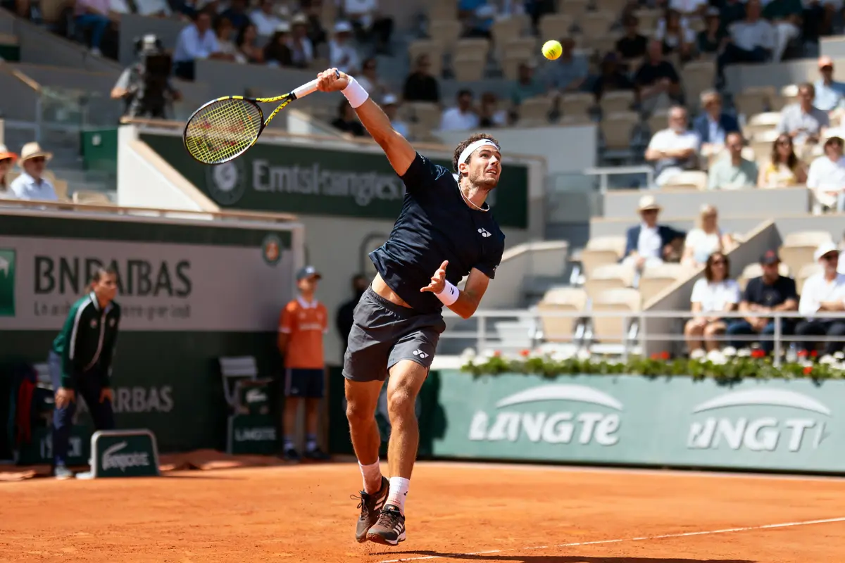 Joueur de tennis en action, frappant la balle sur un court en terre battue, public en arrière-plan.