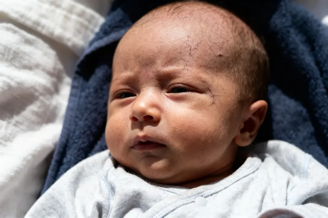 Un bébé allongé, habillé de gris, le visage éclairé par le soleil, expression sérieuse.