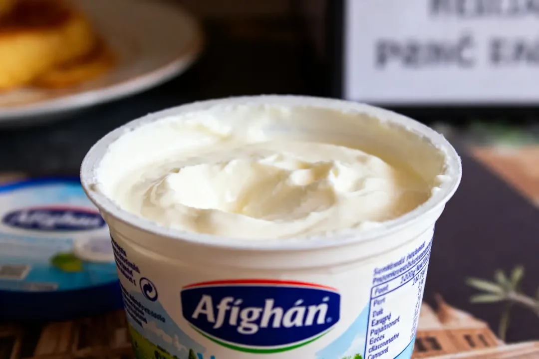 Pot de crème épaisse Afighán ouvert, avec cuillère visible en arrière-plan. Fond flou avec texte.